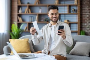Indian young man working from home with laptop and bills, holding receipt and looking smilingly at mobile phone screen, checking documents.